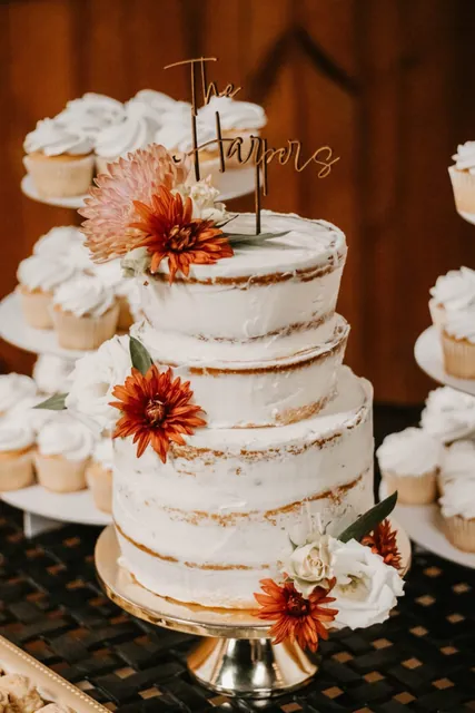 A naked tiered wedding cake with dahlias, cupcakes around it